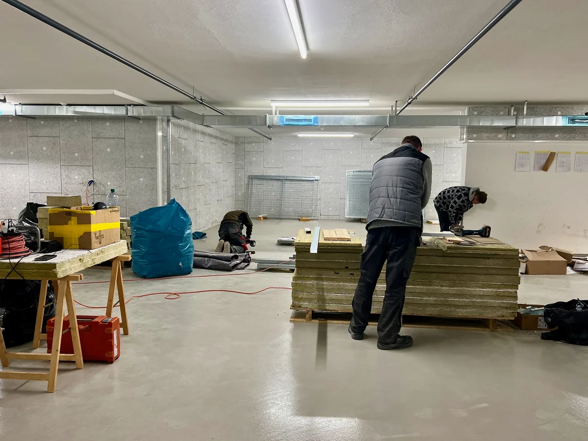 Man sieht drei Arbeiter in einer Tiefgarage, am Schrauben und Zuschneiden von Dämmmaterial.